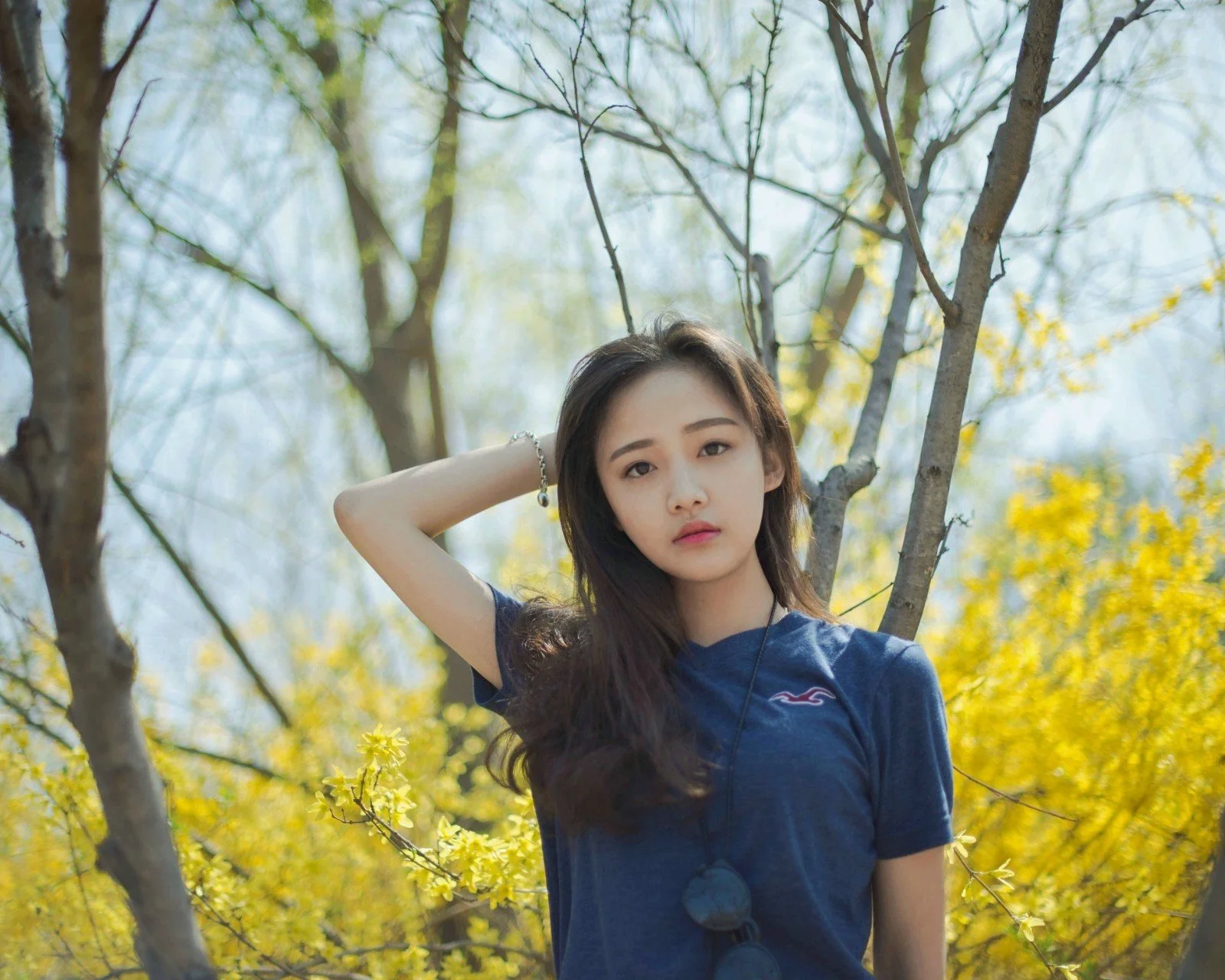 한 젊은 여성이 나무와 노란 꽃 사이에서 한 손으로 긴 머리를 다듬고 있다.