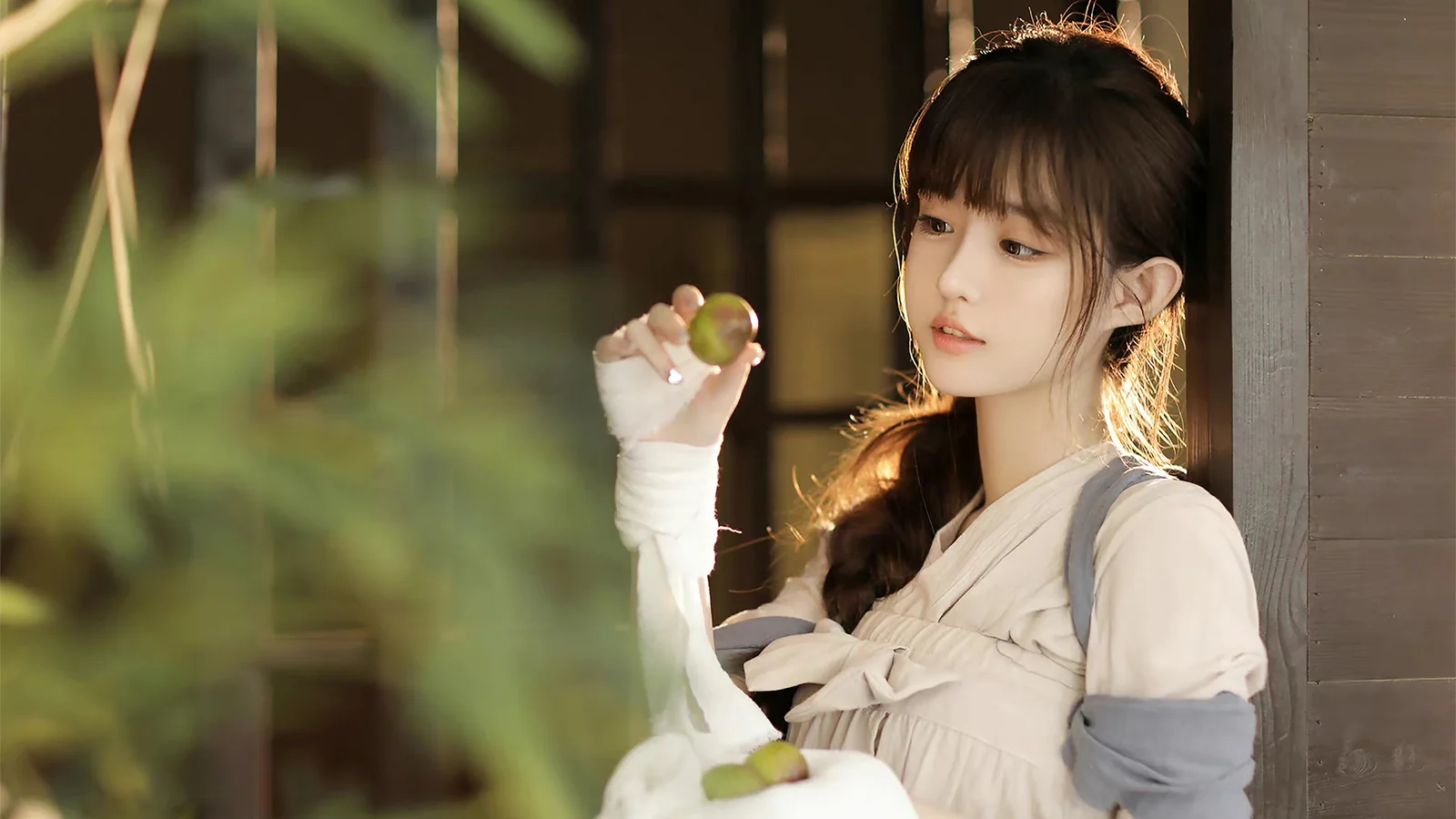 밝은 색상의 옷을 입은 여성이 나무 창가에서 초록색 과일을 들고 따뜻한 햇빛에 윤곽을 드러내고 있다.
