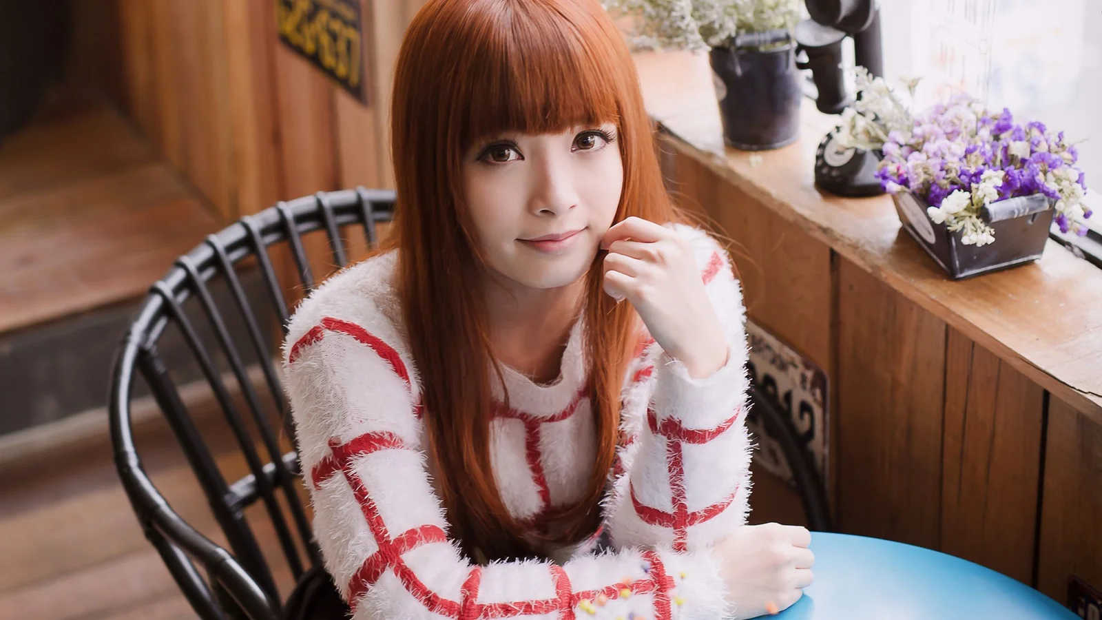 빨간 머리의 여성이 흰색과 빨간색 무늬 스웨터를 입고 카페 창가에서 카메라를 바라보고 있습니다.