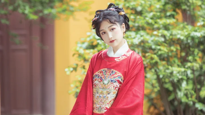 一名女子身穿带有精美刺绣的红色传统风格服饰，站在绿植环绕的庭院中。