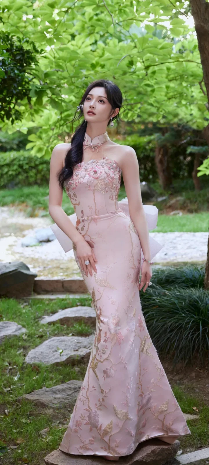 一名成年女性身穿粉色长礼服站在绿意盎然的花园石径旁。