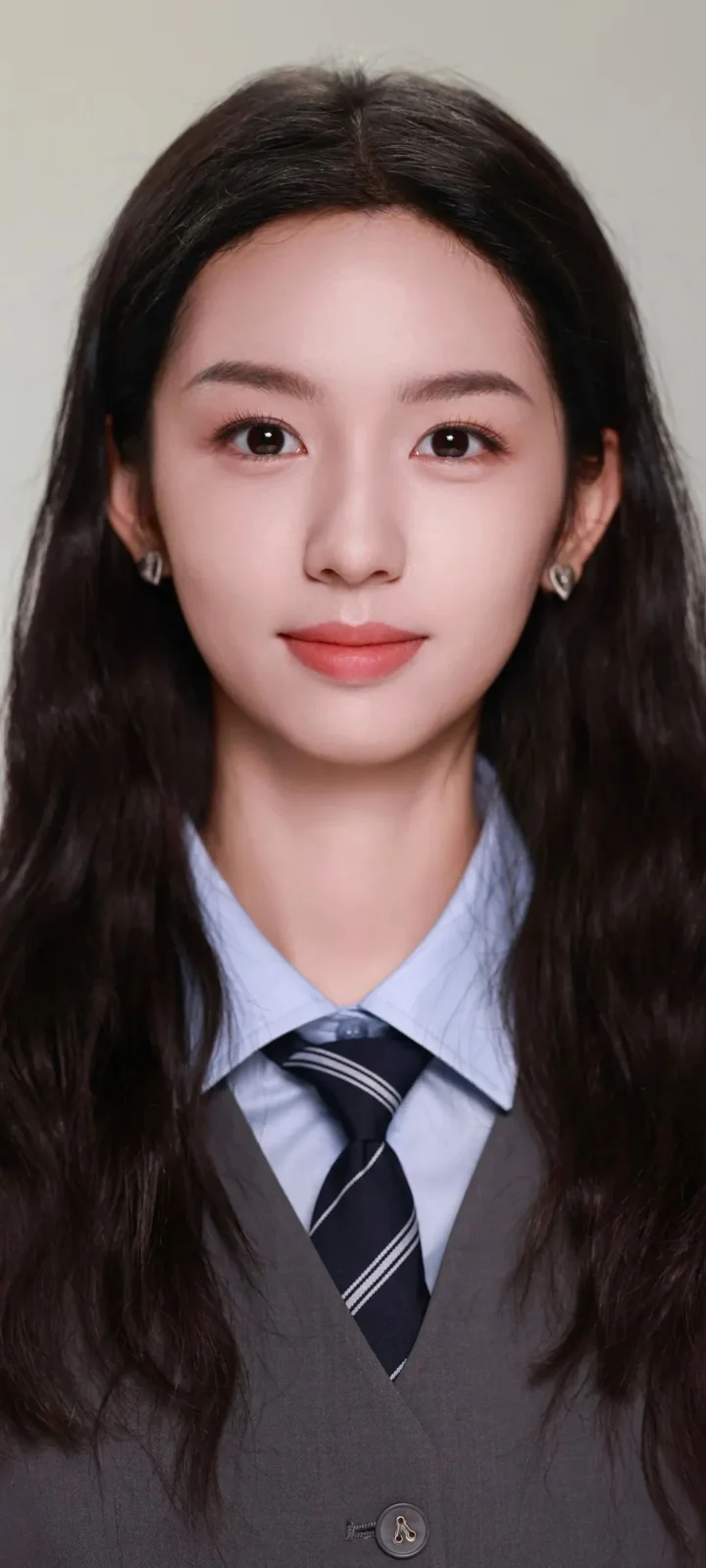 一名长发女性身穿灰色西装外套、浅蓝色衬衫和条纹领带，面向镜头站在浅色背景前。