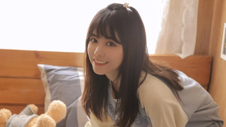 一名长发女子坐在卧室的床上微笑面对镜头。