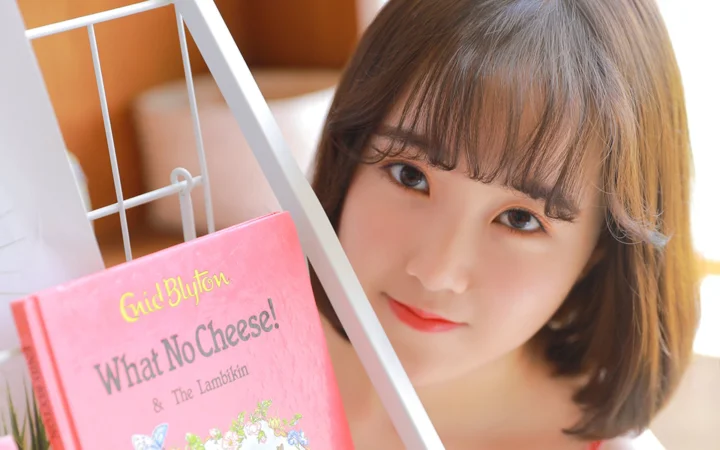 갈색 단발머리와 앞머리를 한 여성이 얼굴 옆의 분홍색 책과 함께 카메라를 바라보고 있습니다.