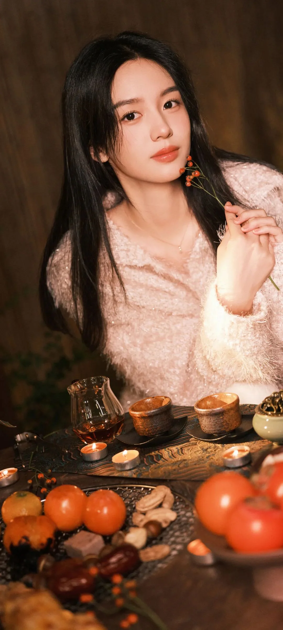 一位成年女性手持带红色果实的小枝条，坐在摆有茶具和水果的桌边。