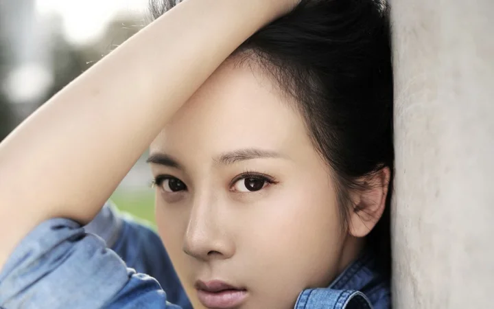 성인 여성이 밝은 색 벽에 기대어 한쪽 팔을 올린 채 파란 데님 상의를 입고 있는 얼굴 중심의 클로즈업입니다.