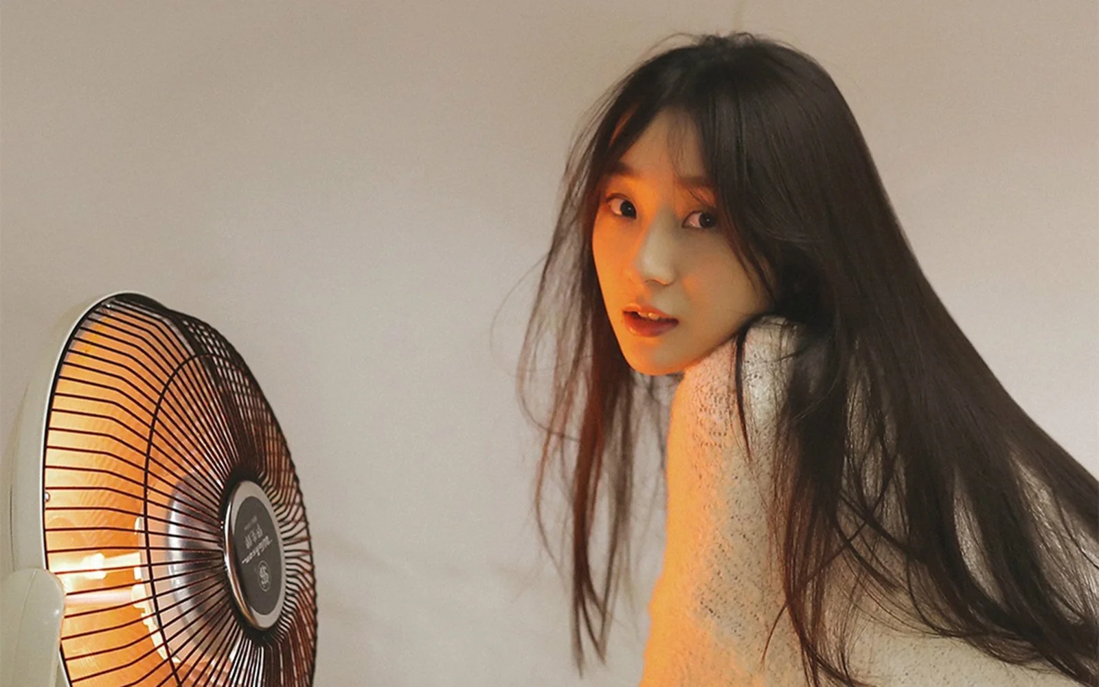 一名长发女子穿着浅色针织衫坐在电风扇旁，暖色灯光映照着室内画面。