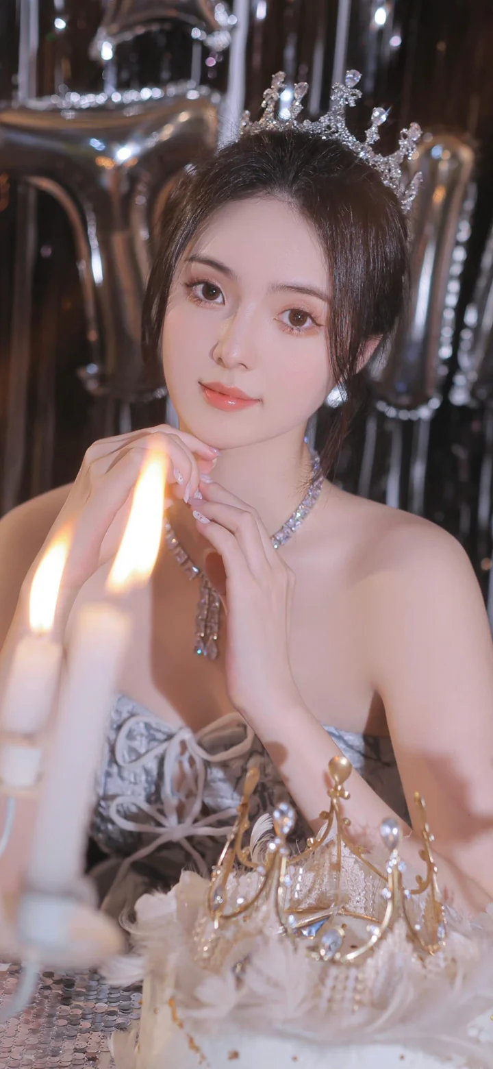 一名戴着王冠和珠宝、身穿浅色礼服的女性面对镜头摆姿势，双手靠近下巴。