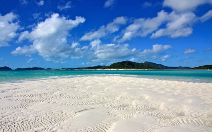 洁白沙滩延伸至碧 turquoise 海水，远处是绿色海岛，湛蓝天空点缀着蓬松白云。