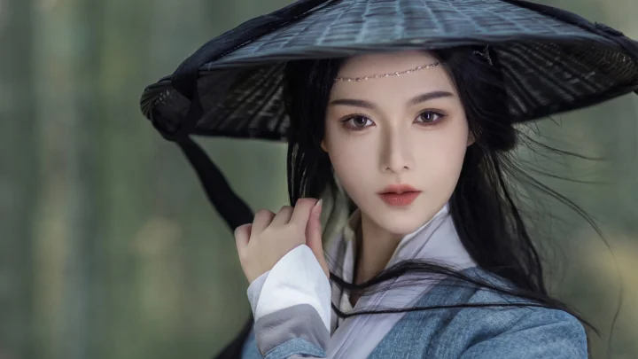 一名戴着宽檐黑色帽子、穿着浅色传统风格服装的成年女性面对镜头。