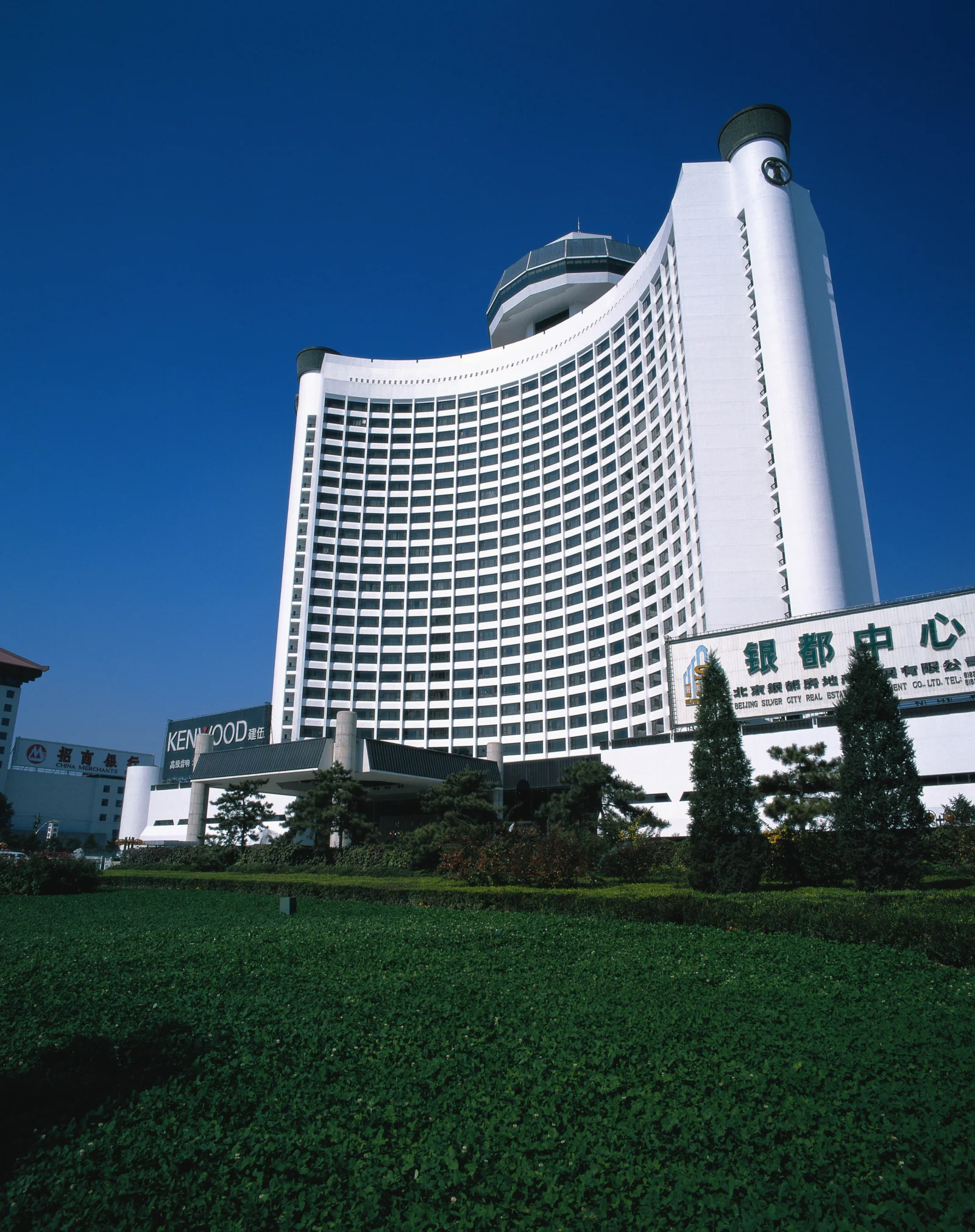 一座白色弧形高层建筑矗立在深蓝天空下，前方是修剪整齐的绿篱。