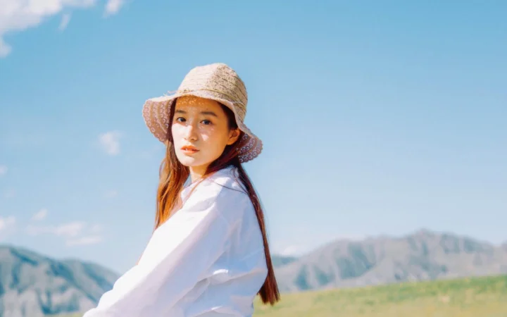 밀짚모자를 쓴 여성이 푸른 하늘 아래 산을 배경으로 서 있습니다.