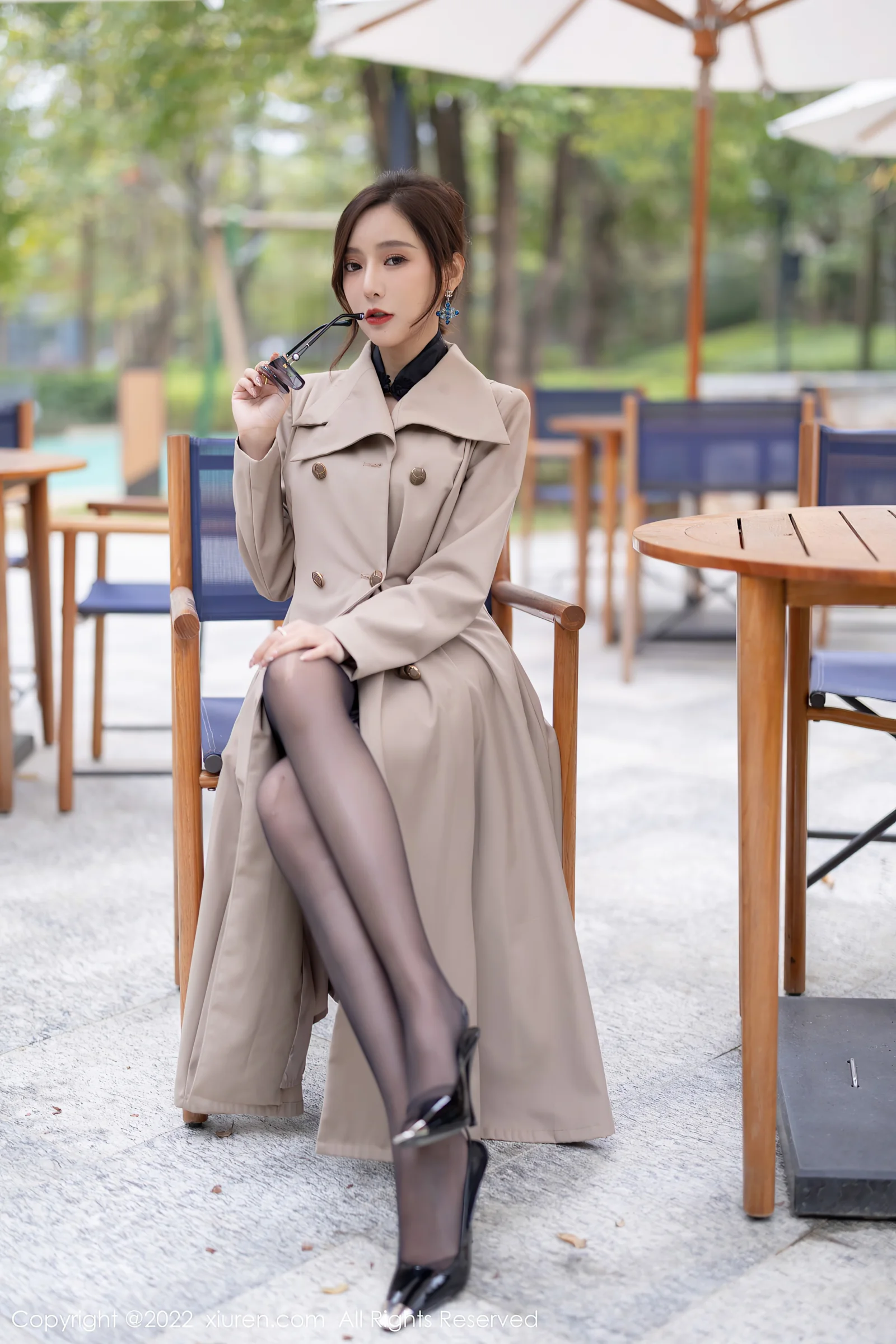 베이지색 트렌치코트를 입은 성인 여성이 선글라스를 손에 들고 야외 카페 의자에 앉아 있습니다.
