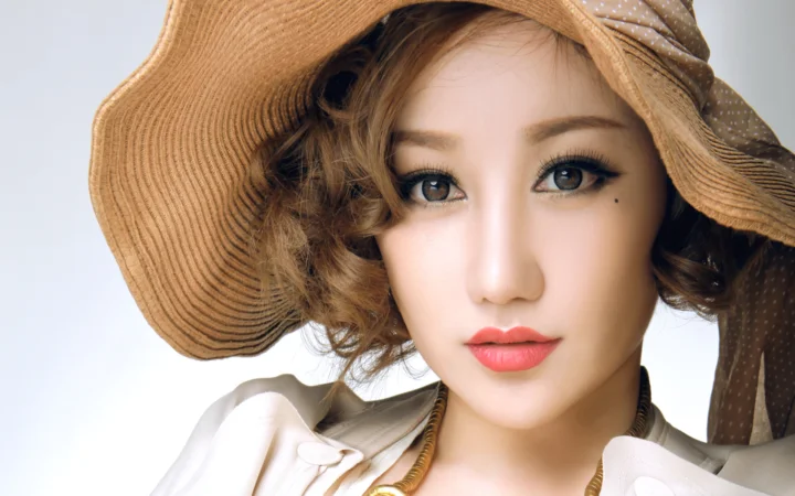 곱슬머리의 여성이 챙이 넓은 밀짚모자와 밝은 색 상의를 입고 카메라를 향해 미소 짓고 있다.