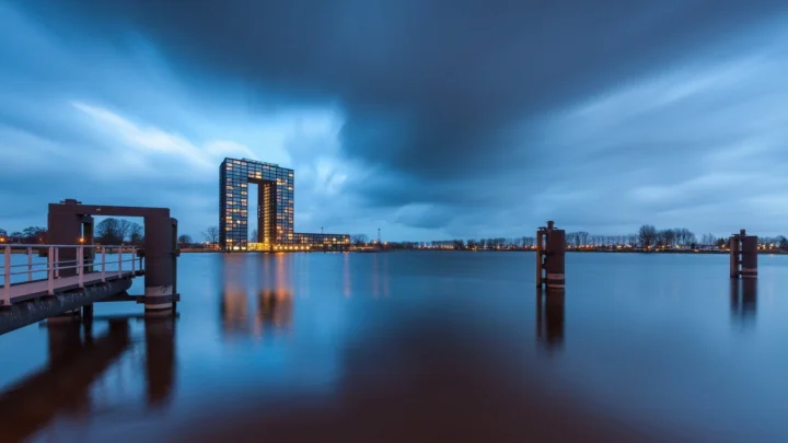 A long-exposure blue hour view of a rectangular high-rise over still water, with a pier, wooden posts, and smooth cloud reflections. Calm 16:9 desktop landscape wallpaper.