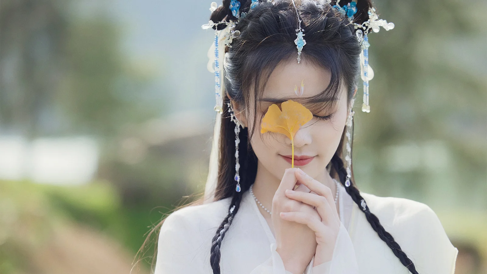 흰색 전통 의상을 입은 여성이 야외에서 눈을 감고 금빛 잎을 들고 있는 부드러운 배경의 인물 사진이다.