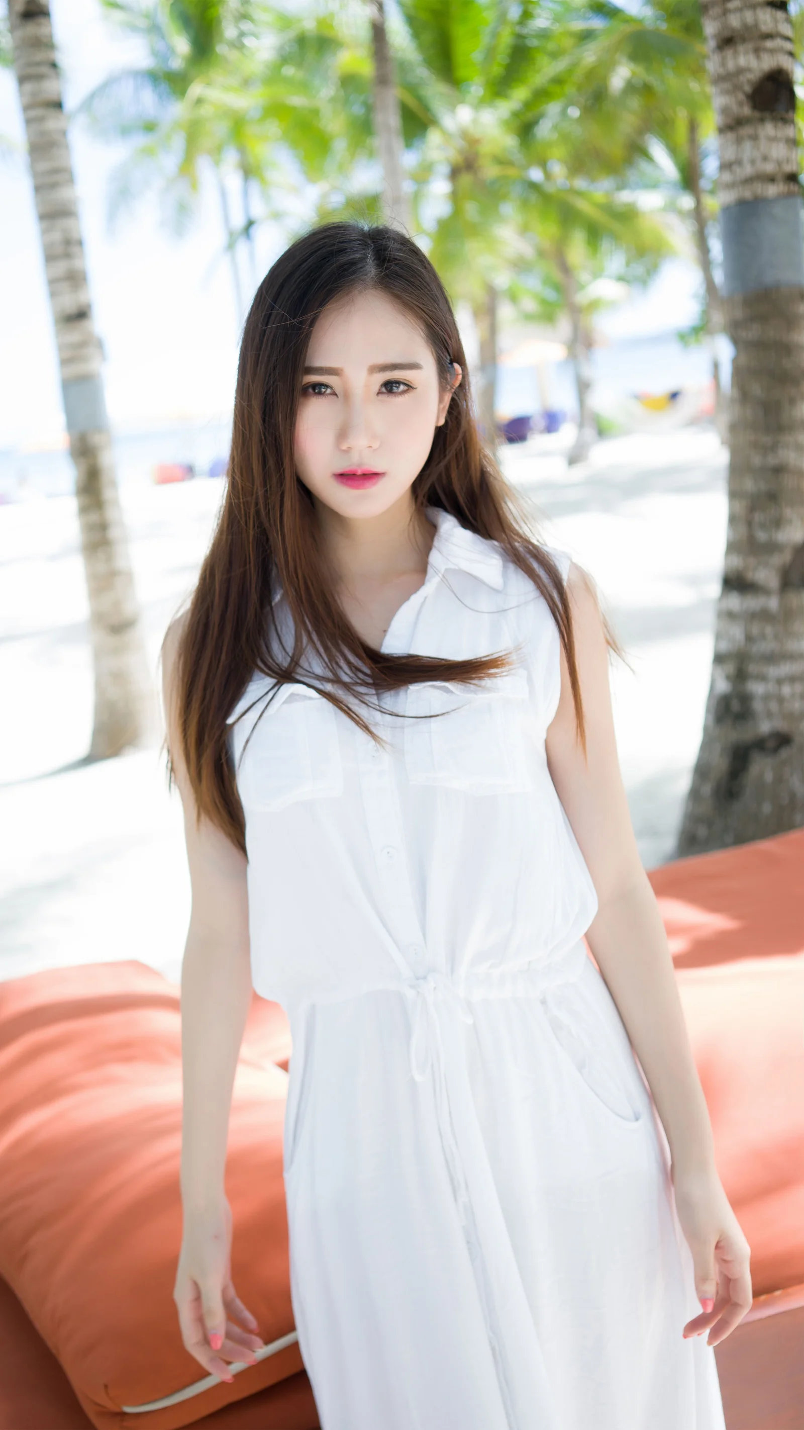 一名身穿白色连衣裙的成年女子站在棕榈树环绕的明亮海滩上。