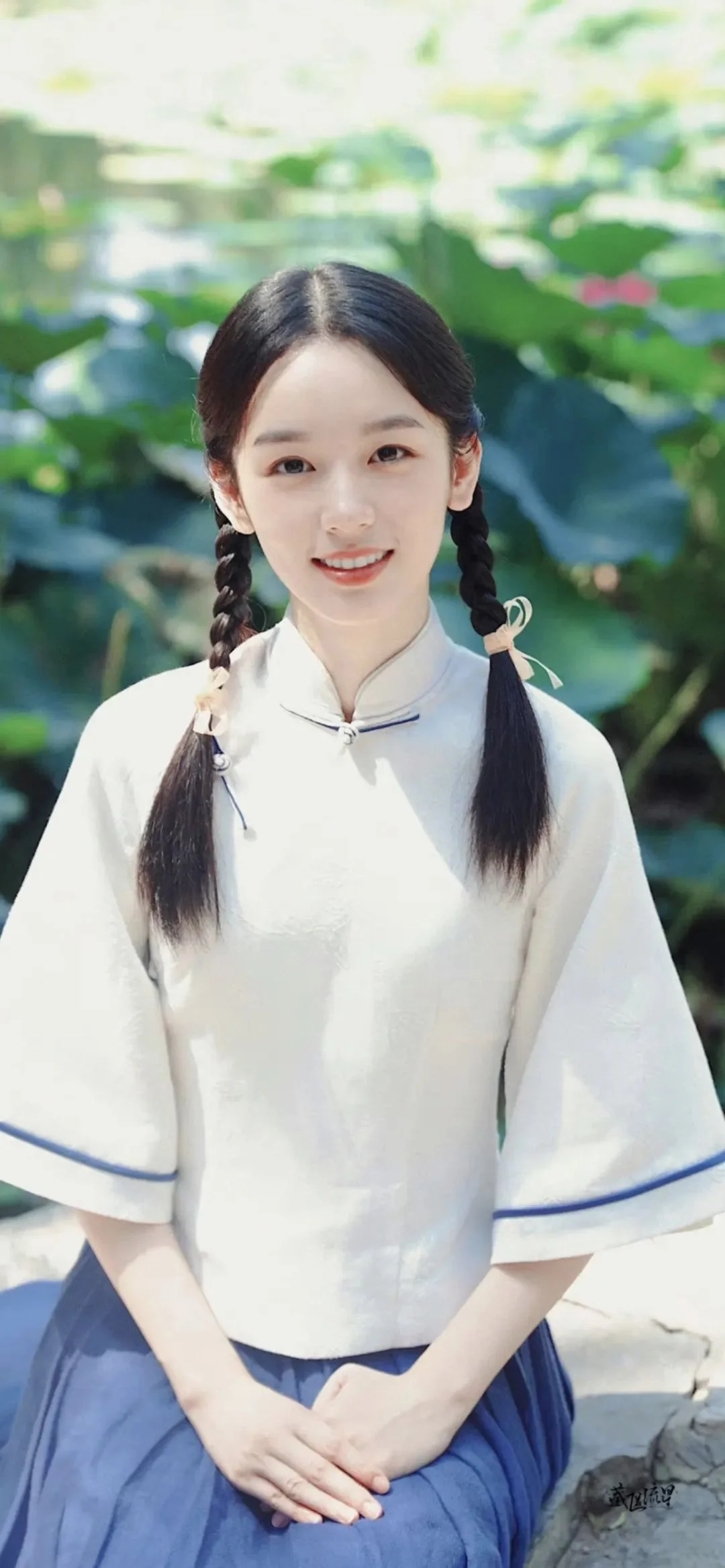 一名扎着双辫、身穿浅色传统服饰的女子站在绿叶背景前。