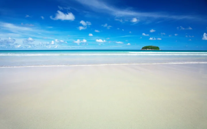 湛蓝天空下的白沙海滩延伸至清澈海水，远处可见一座绿色小岛。