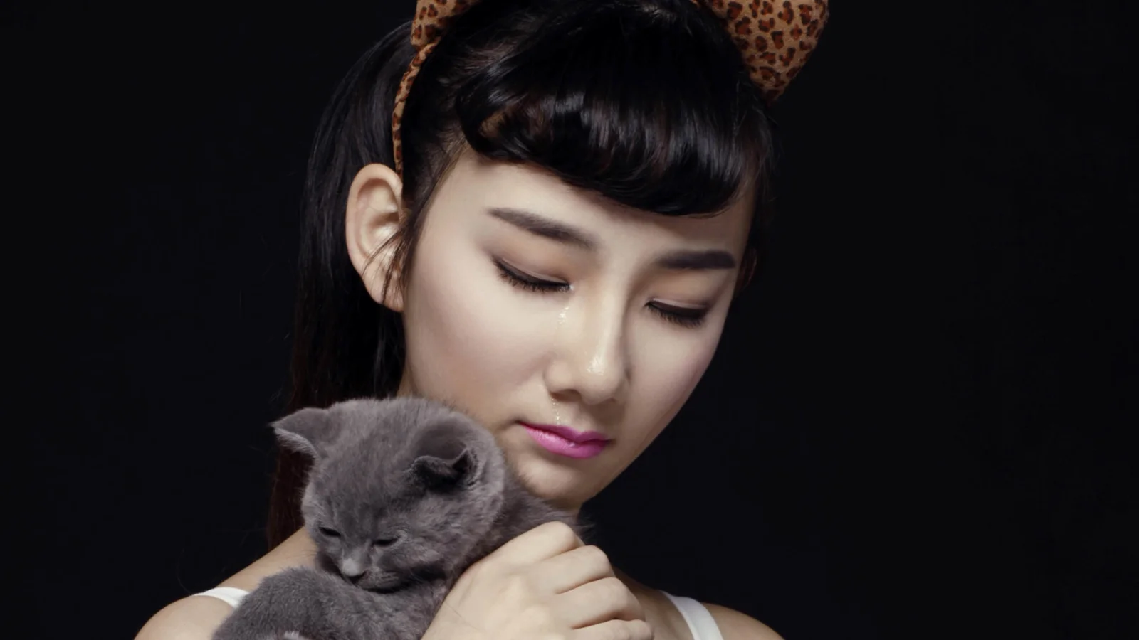 黑色背景前，一名闭眼的女子抱着一只灰色小猫。