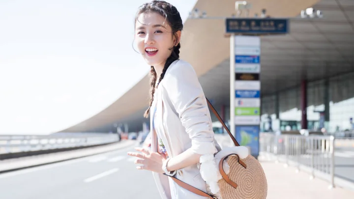 一名身穿白色外套、背着圆形编织包的女子站在机场航站楼旁微笑。