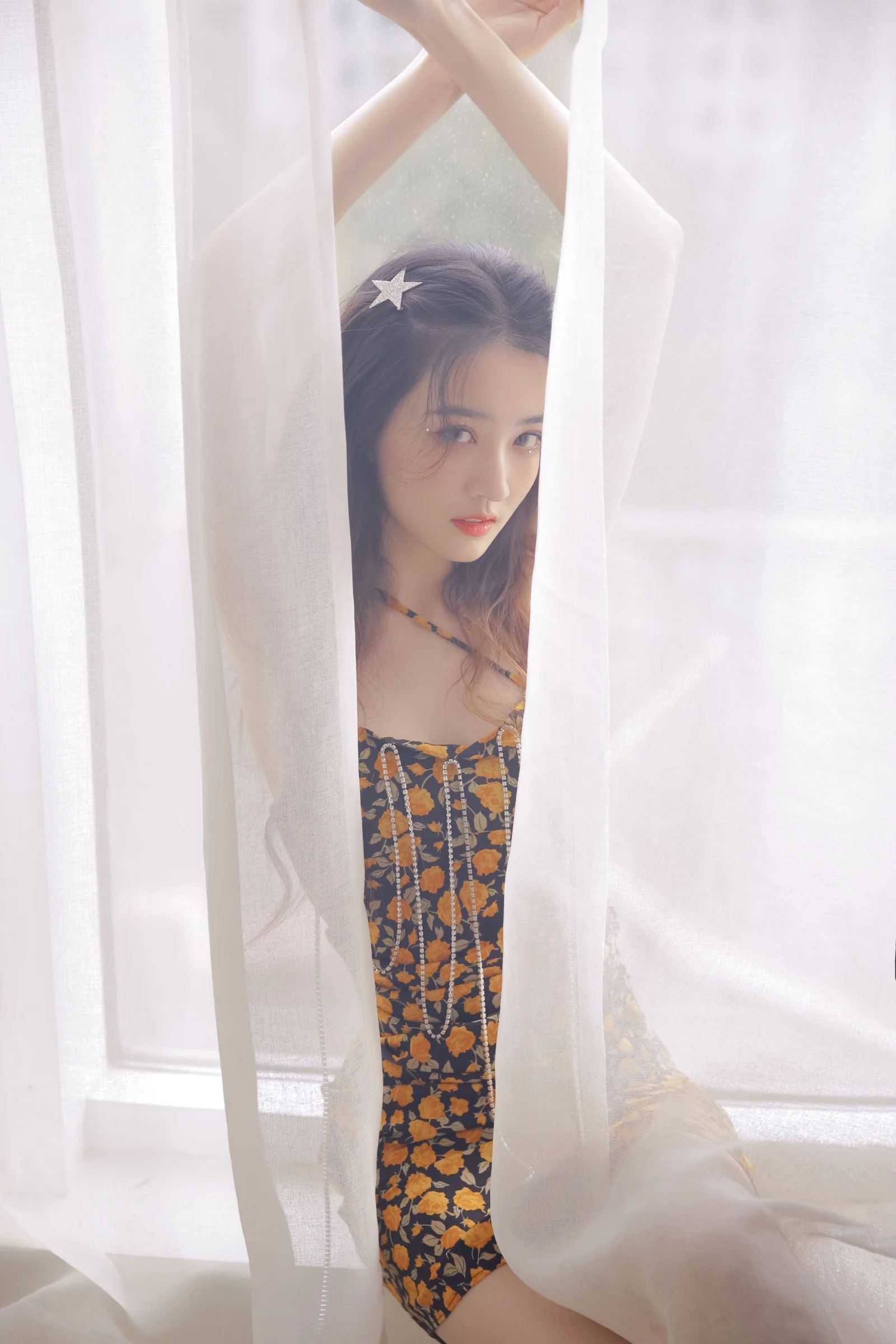 一名身穿花卉图案连衣裙的女性站在白色纱帘之间，窗边柔光映照着她的脸庞。