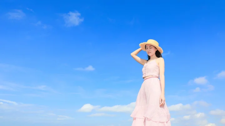 一名戴草帽、穿粉色长裙的女性站在蓝天白云下。