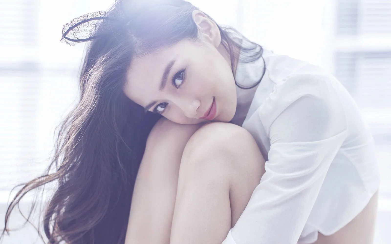 一名长发女子穿着白色长袖上衣，侧头靠在膝盖上面对镜头。