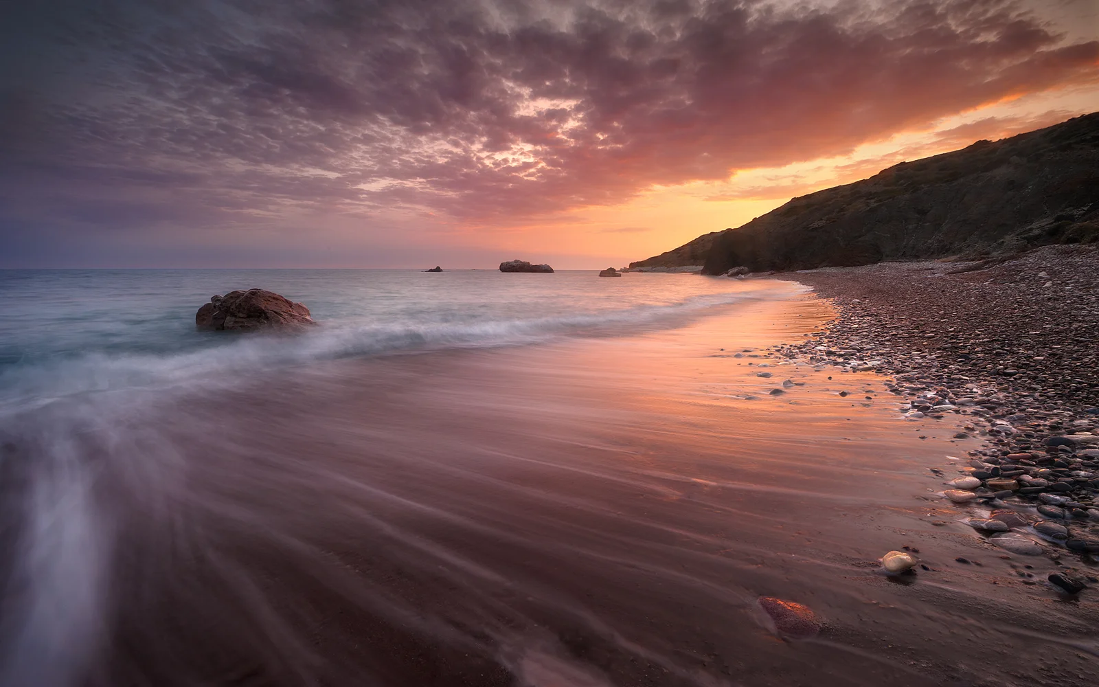 夕阳映照下的海岸沙滩延伸至远处的岩石海岬，海水在岸边缓缓流动。