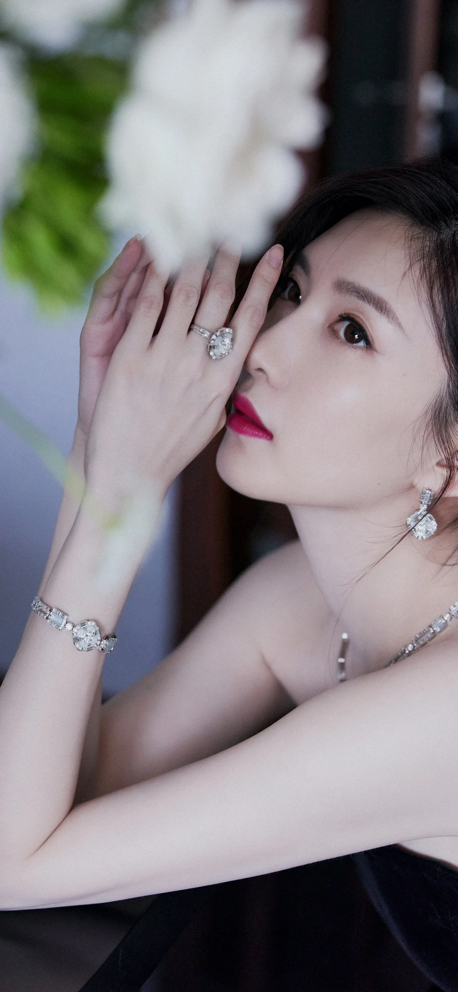 一位成年女性以手靠近脸部的姿态面对镜头，佩戴耳环、戒指、手链和项链，背景虚化。