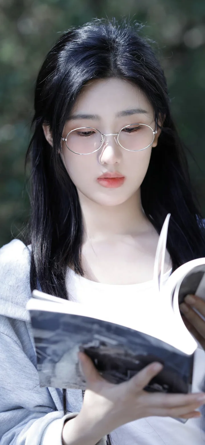 一名戴圆框眼镜的女子手持书本站在虚化的绿色背景前。