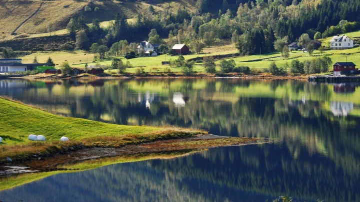 宁静湖泊倒映着森林、山坡与湖畔乡村房屋，岸边绿地延伸至前景。