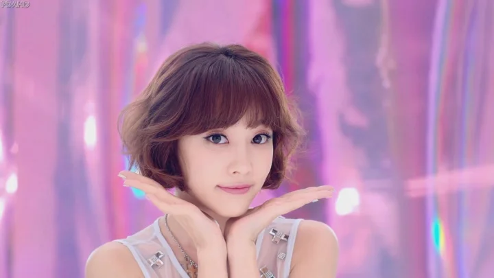 단발머리 여성이 분홍색과 보라색 조명을 배경으로 턱을 손에 괴고 미소 짓고 있다.