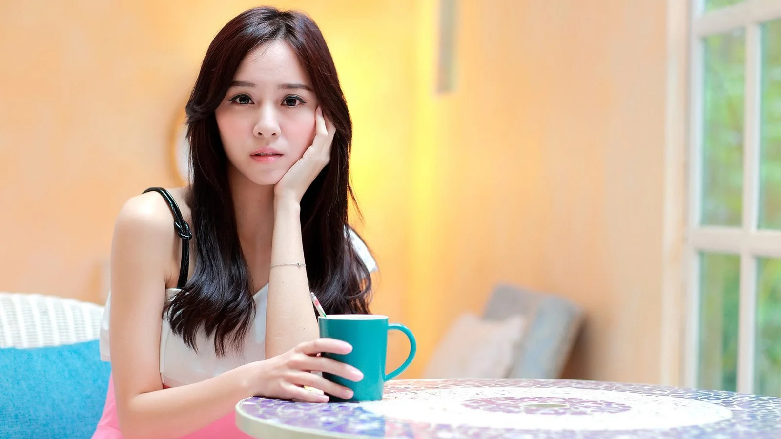 성인 여성이 실내 창가에 앉아 청록색 머그컵을 들고 카메라를 바라보고 있다.