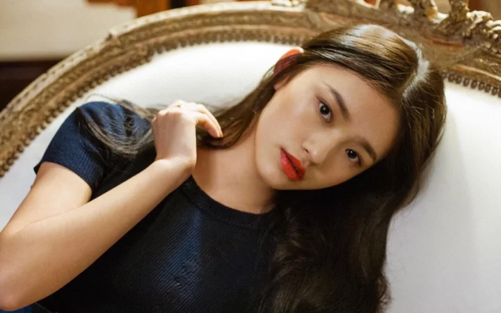 검은 머리의 여성이 장식 의자에 기대어 카메라를 바라보고 있습니다.