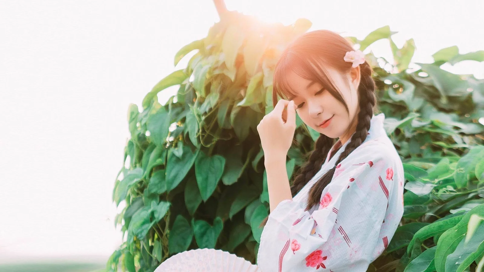柔らかな日差しの中、緑の葉と花を背景に成人女性が立っています。