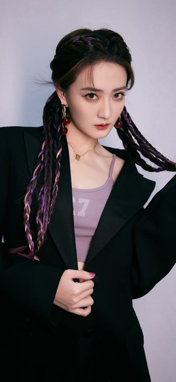 컬러풀한 땋은 머리의 여성이 검은색 블레이저를 입고 밝은 배경 앞에서 포즈를 취하고 있다.