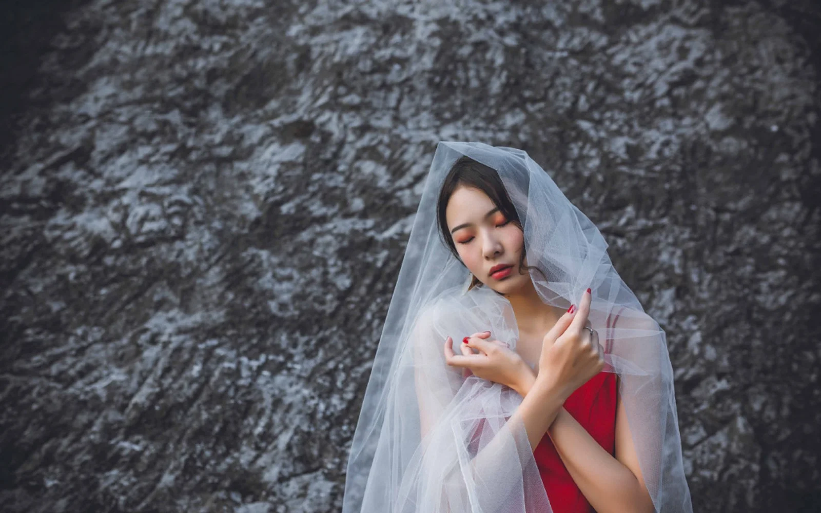 一名成年女性闭着眼睛，身披半透明薄纱并穿着红色服装，站在岩石墙前。