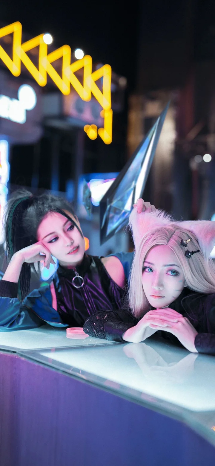 2人の成人女性コスプレイヤーが、ネオンライトに照らされながら光るテーブルのそばに寄り添っている。