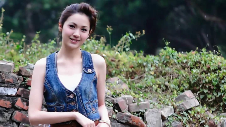 一名身穿牛仔马甲的女性站在绿植和石墙前微笑。