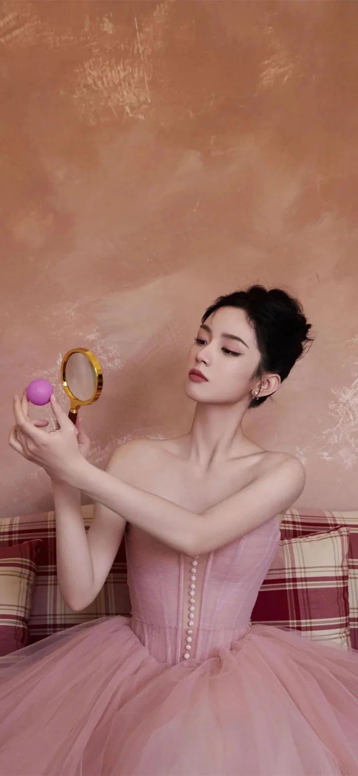 一名成年女性身穿粉色礼服，手持小镜子和紫色圆球坐在格纹沙发前。