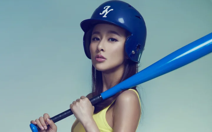 一名戴着蓝色棒球头盔、手持蓝色球棒的女性站在浅蓝色背景前。