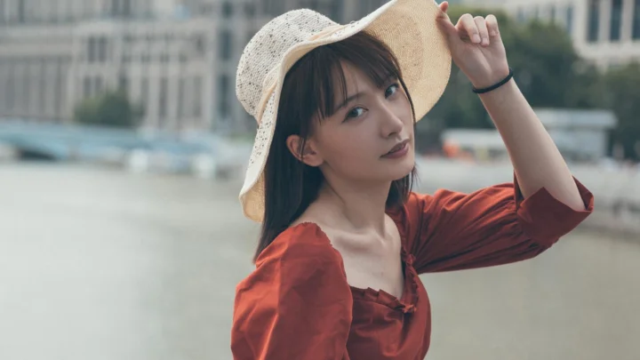 밀짚모자와 빨간 원피스를 입은 여성이 야외 물가에 서 있다.