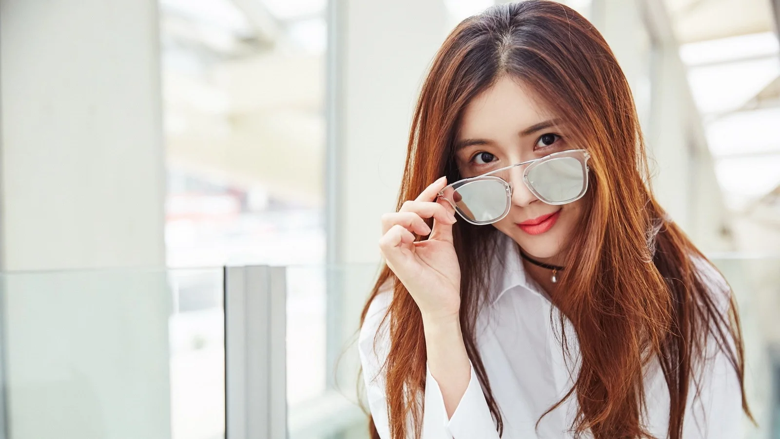 긴 갈색 머리의 여성이 흰 셔츠를 입고 밝은 실내 창가에서 선글라스를 든 채 카메라를 바라보고 있다.