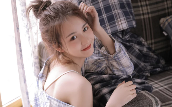 一名成年女性靠在格纹床品旁回望镜头，窗边柔和的光线照亮了她的脸庞。