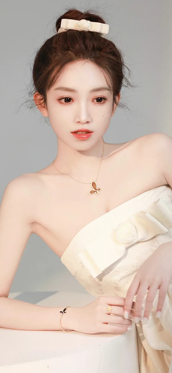 一位盘发女性身穿浅色无肩带礼服，面对镜头坐在浅灰色背景前。