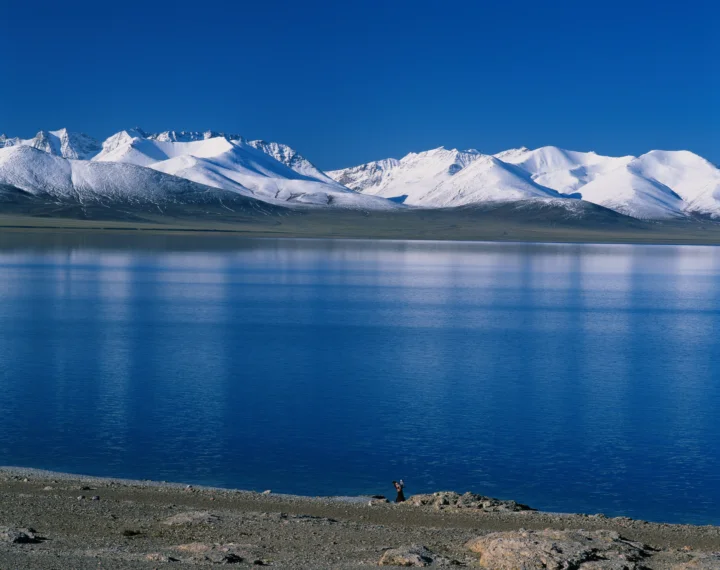 蓝色湖泊对岸绵延着覆雪山脉，前景是荒凉的岩石湖岸。