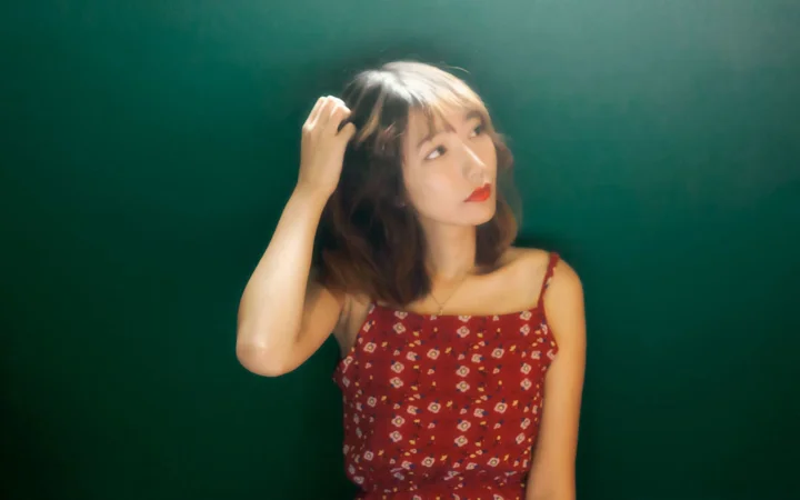 一名成年女性身穿红色碎花吊带裙，在深绿色背景前轻抚头发并望向一侧。