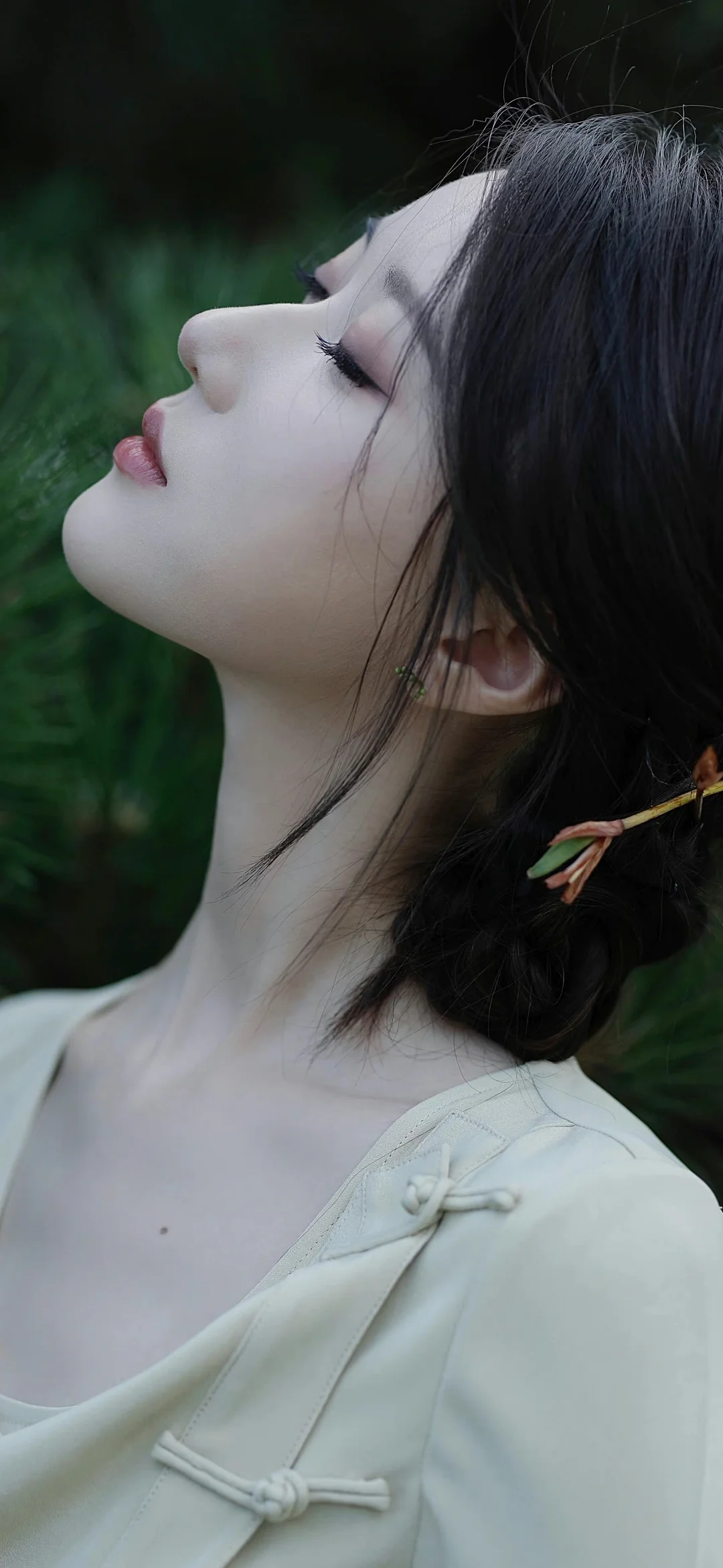 一位黑发女性身穿白色衣服，在绿色植物背景前呈侧脸姿态。