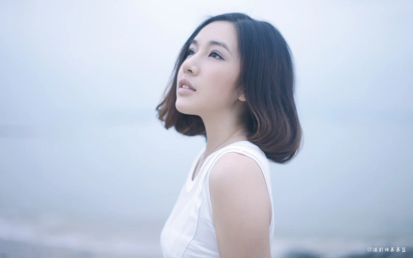 白いノースリーブを着た成人女性が、淡い青色の背景の前で遠くを見上げています。